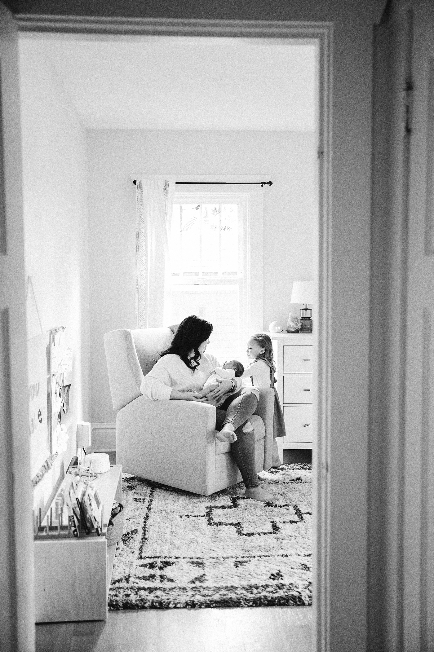 Mom rocks baby boy in chair while talking to her oldest daughter. Photo by Meg Newton Photography.