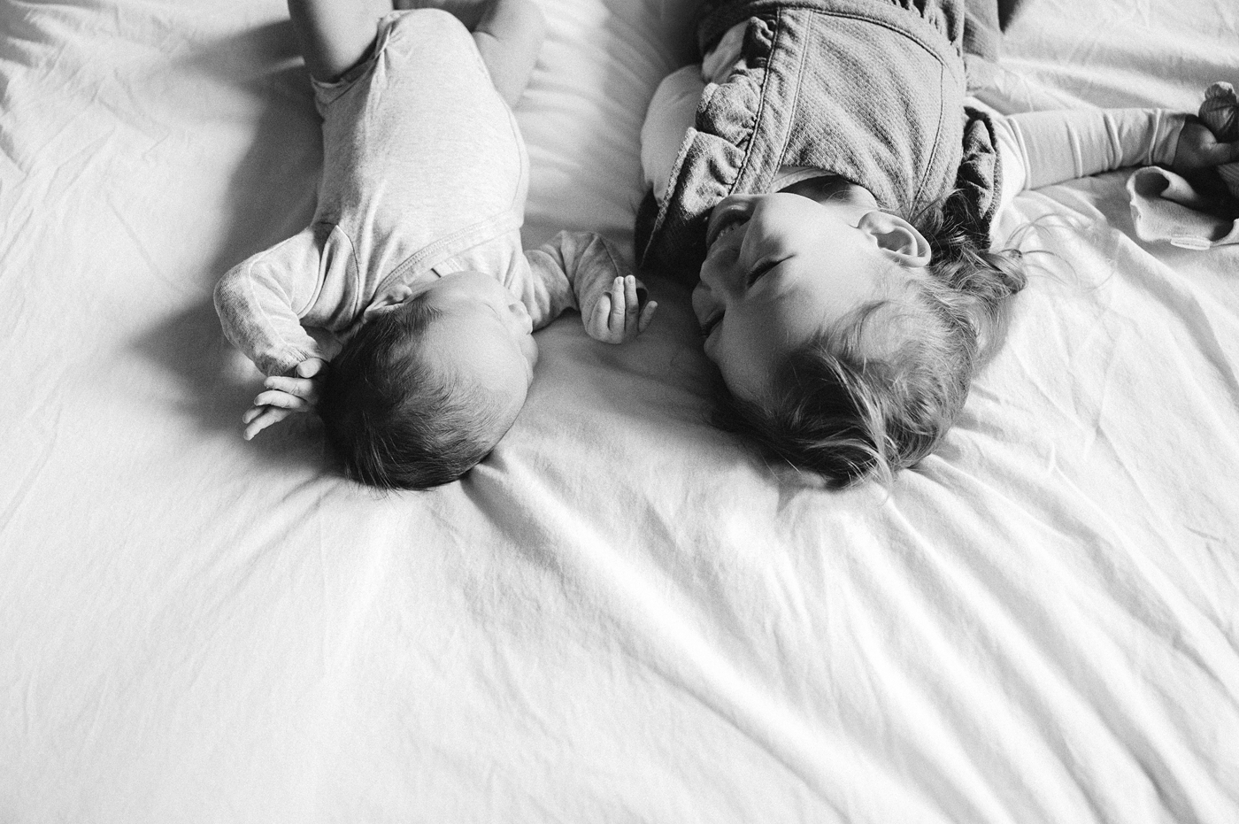 Sister giggles at baby brother while laying on back in bed. Photo by Meg Newton Photography.