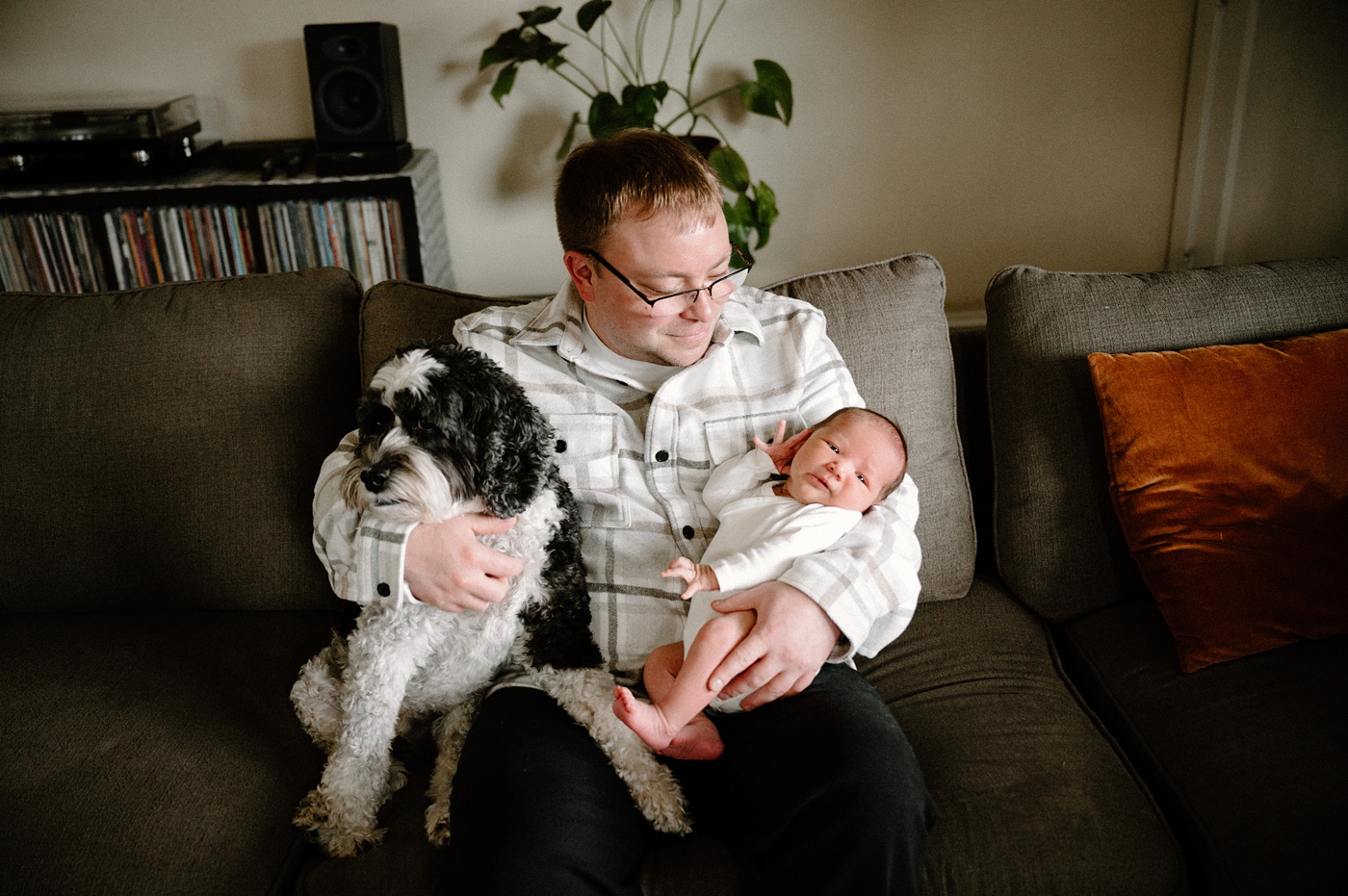 Dad holds his dog and newborn son while sitting on a gray couch. Photo by Meg Newton Photography.