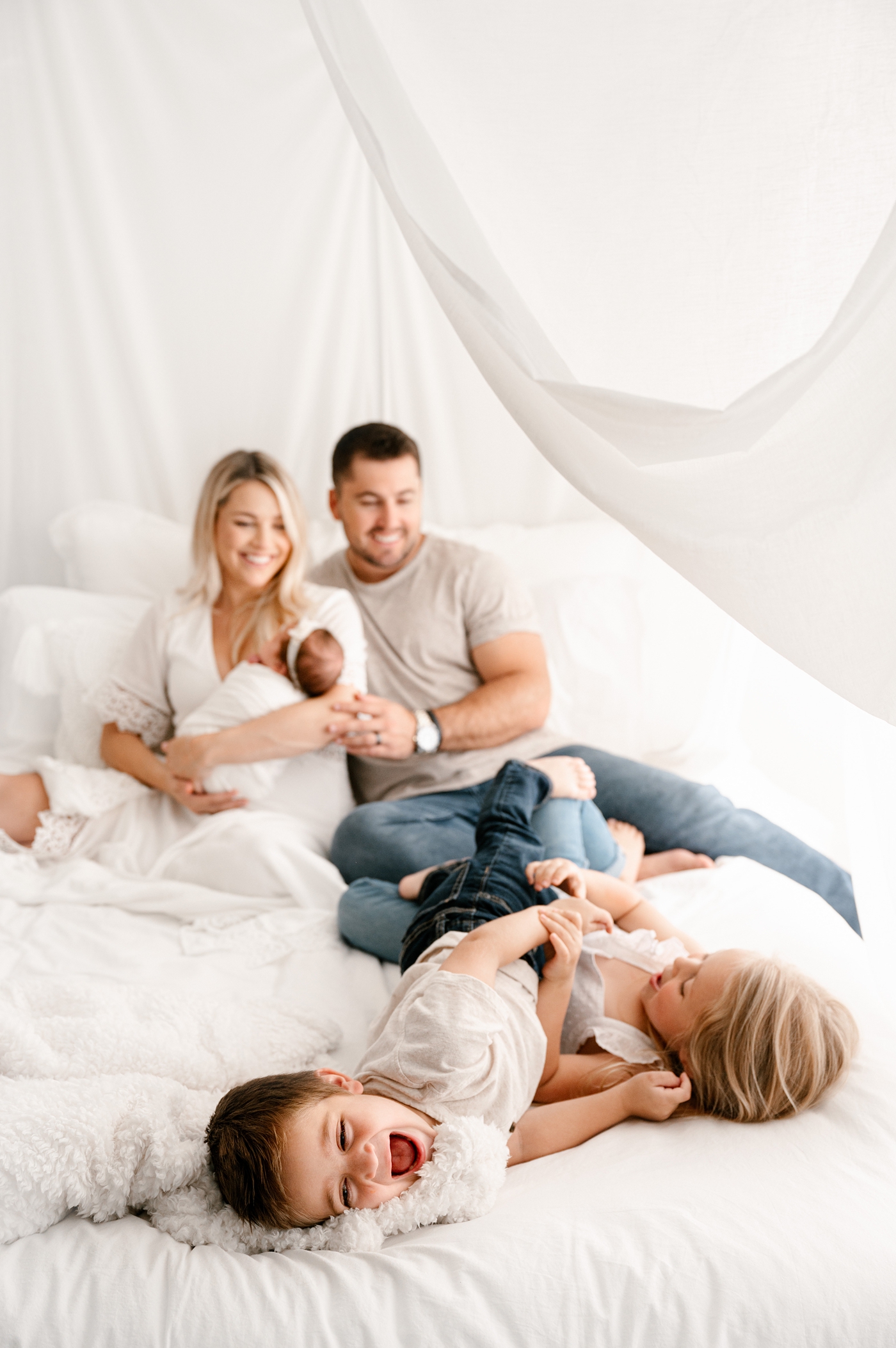Children wrestle on the end of bed during lifestyle studio newborn session. Image by Meg Newton Photography.