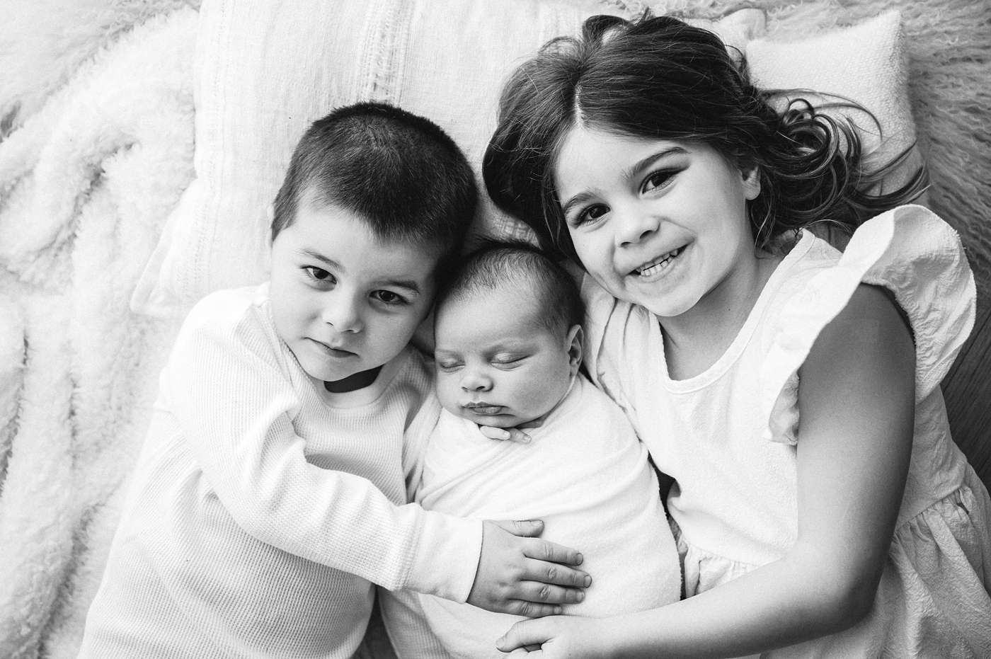Big brother and sister hold baby brother while lying on a bed. Photo by Meg Newton Photography.