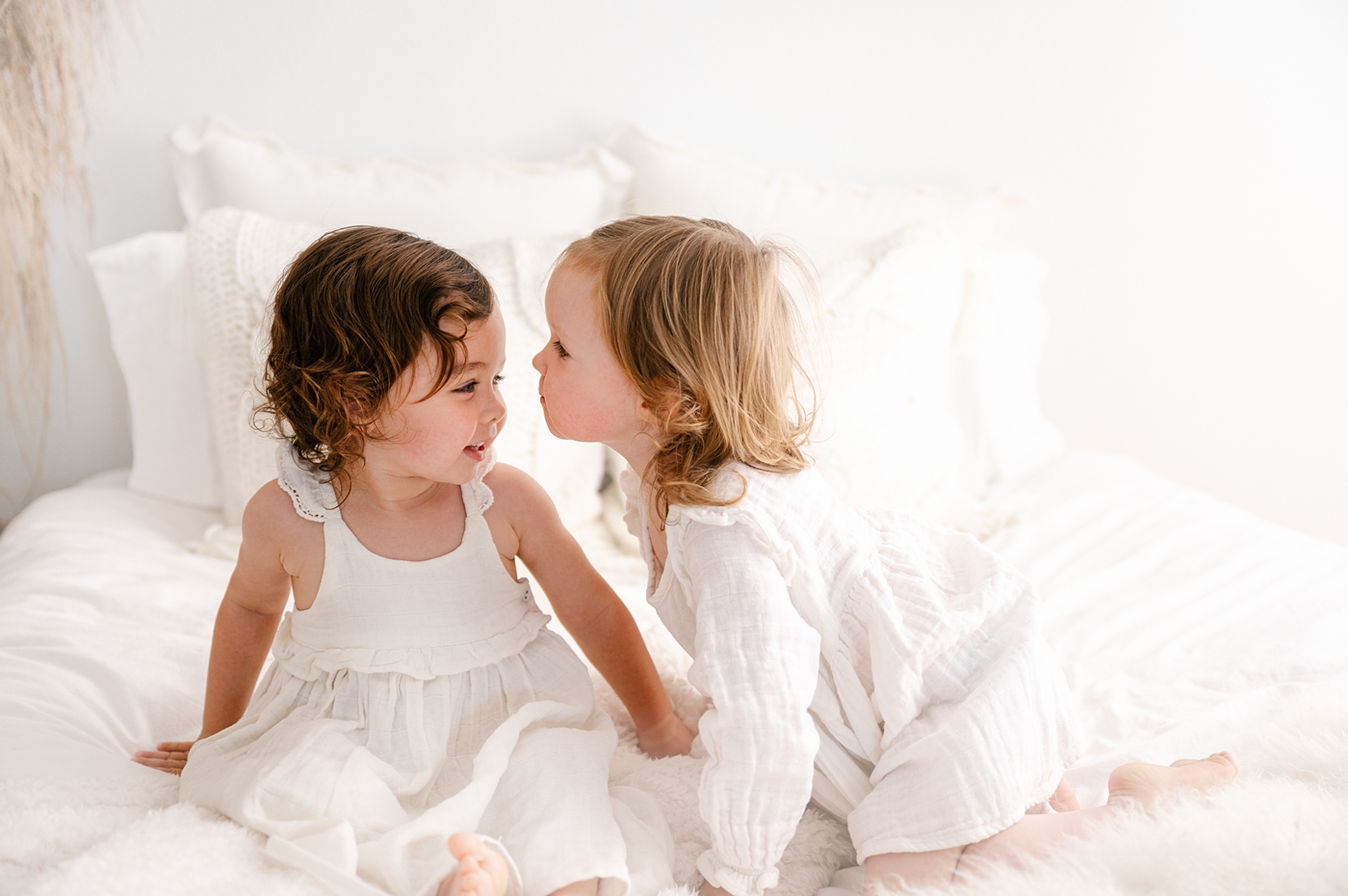 Sisters sitting on all white studio bed together during toddler milestone session. Photo by Meg Newton Photography.