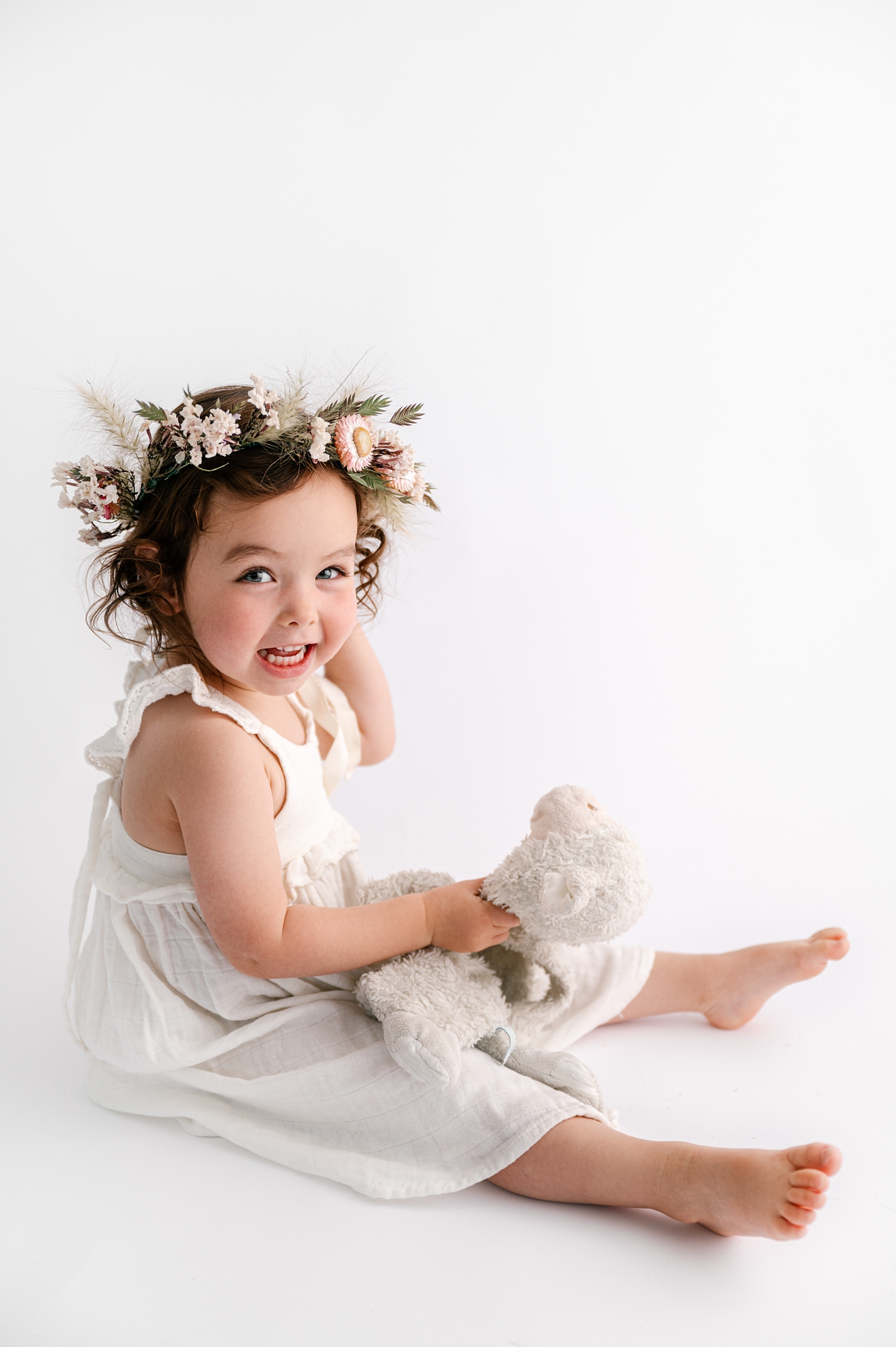 Little girl smiles holding lamb stuffed animal while wearing floral crown. Photo by Meg Newton Photography.